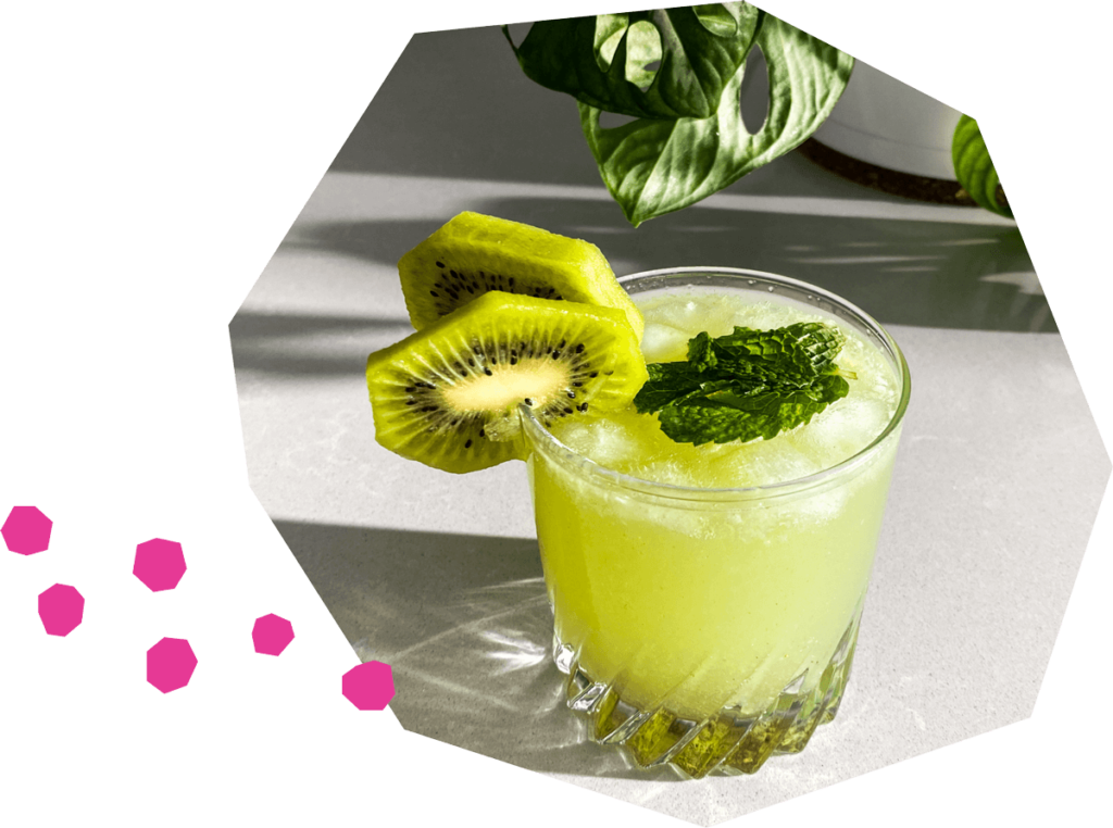 Un verre à cocktail rempli d'eau pétillante jaune-vert pâle, garni de glaçons, de feuilles de menthe et de rondelles de kiwi. Le verre repose sur un comptoir blanc, à côté d'une plante verte en pot.