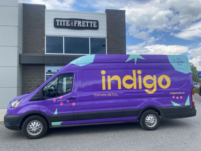 Une camionnette de livraison Indigo, de couleur mauve et décorée de motifs abstraits, est garée devant un magasin Tite Frette. Le logo d'Indigo et la mention "Cylindre de CO2" sont visibles sur le côté de la camionnette.